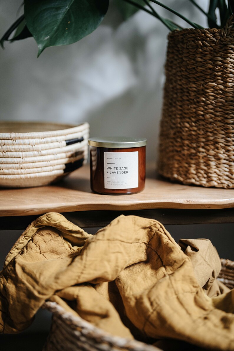 May include: A brown glass candle with a gold lid and a white label that reads "White Sage + Lavender". The candle is sitting on a wooden surface with a woven basket and a yellow blanket.