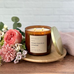 May include: A brown glass candle jar with a gold lid. The candle is labeled "WICKRY CANDLE CO HONEYSUCKLE JASMINE 100% NATURAL SOY WAX 17 OZ | BURN TIME 40+ HRS". The candle is sitting on a wooden coaster and surrounded by pink flowers.