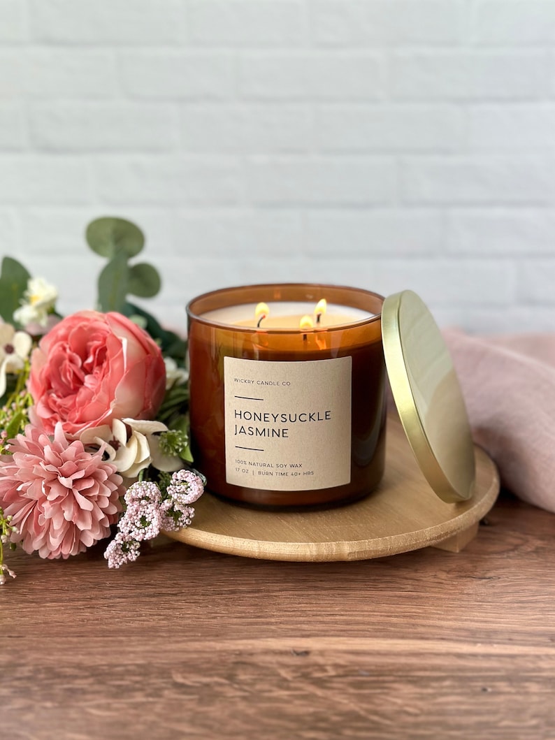May include: A brown glass candle jar with a gold lid. The candle is labeled "WICKRY CANDLE CO HONEYSUCKLE JASMINE 100% NATURAL SOY WAX 17 OZ | BURN TIME 40+ HRS". The candle is sitting on a wooden coaster and surrounded by pink flowers.