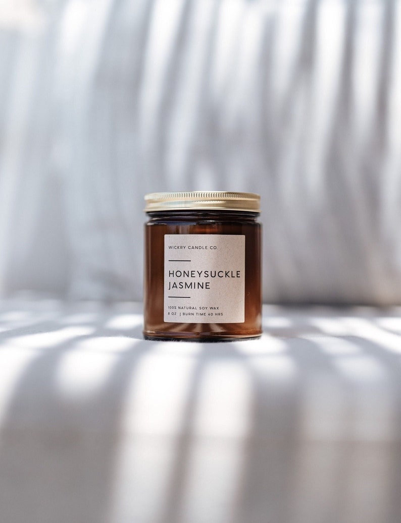May include: A brown glass candle jar with a gold lid. The label reads "WICKRY CANDLE CO. HONEYSUCKLE JASMINE 100% NATURAL SOY WAX 8 OZ | BURN TIME 40 HRS".
