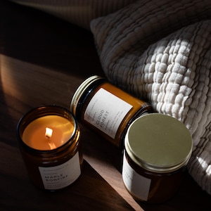 May include: Three amber-colored glass jar candles with gold lids and white labels. One candle is lit, showing a warm glow. The labels read "Maple Bonfire" and "Cloud Forest". The candles are on a dark wooden surface.