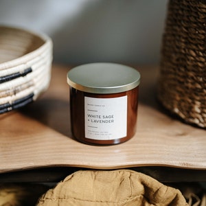 May include: A brown glass candle jar with a gold lid. The label reads "Wickery Candle Co. White Sage + Lavender. 100% Natural Soy Wax. 14 oz | Burn Time 70+ hrs."