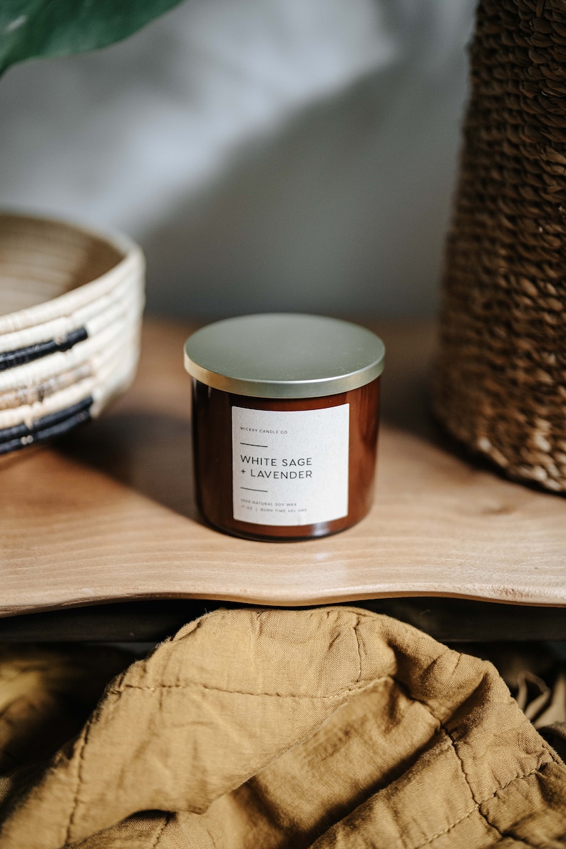 May include: A brown glass candle jar with a gold lid. The label reads "Wickery Candle Co. White Sage + Lavender. 100% Natural Soy Wax. 14 oz | Burn Time 70+ hrs."
