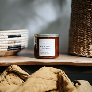 May include: A brown glass candle jar with a gold lid. The label reads "White Sage + Lavender" and "100% Natural Soy Wax 4.2 oz | Burn Time 40-50 Hours".