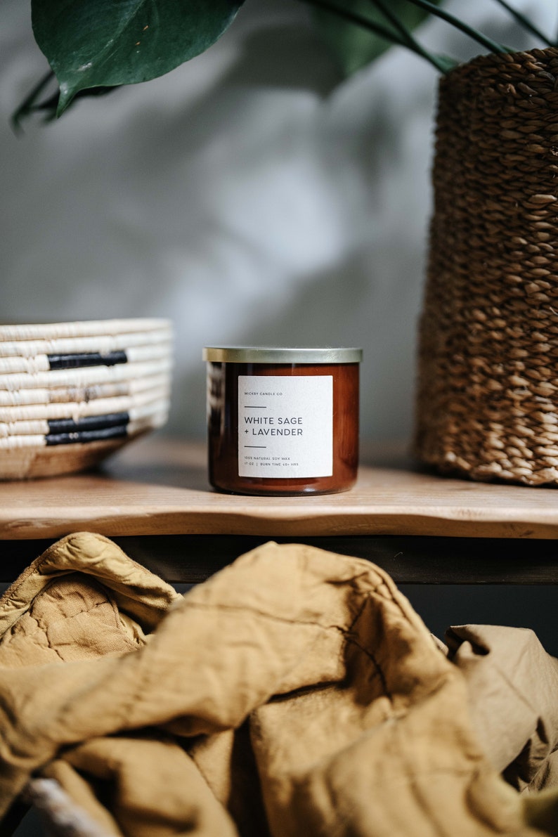 May include: A brown glass candle jar with a gold lid. The label reads "White Sage + Lavender" and "100% Natural Soy Wax 4.2 oz | Burn Time 40-50 Hours".