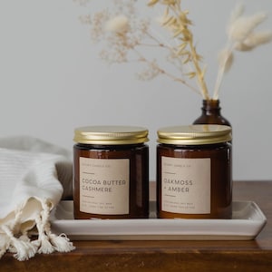 May include: Two amber glass candle jars with gold lids and brown paper labels. The labels read "Cocoa Butter Cashmere" and "Oakmoss + Amber". The candles are on a white rectangular tray on a wooden surface.
