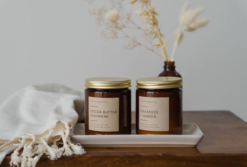 May include: Two amber glass candle jars with gold lids and brown paper labels. The labels read "Cocoa Butter Cashmere" and "Oakmoss + Amber". The candles are on a white rectangular tray on a wooden surface.