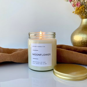 May include: A clear glass candle jar with a gold lid. The candle is labeled "WICKRY CANDLE CO. MOONFLOWER 100% NATURAL SOY WAX 8 OZ | BURN TIME 40 HRS".