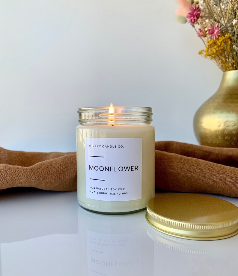 May include: A clear glass candle jar with a gold lid. The candle is labeled "WICKRY CANDLE CO. MOONFLOWER 100% NATURAL SOY WAX 8 OZ | BURN TIME 40 HRS".