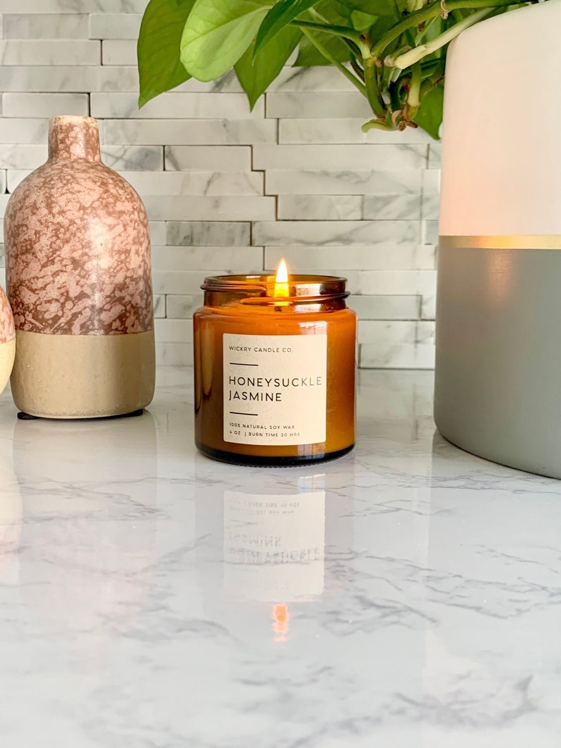 May include: A brown glass candle with a white label that reads "WICKERY CANDLE CO. HONEYSUCKLE JASMINE 100% NATURAL SOY WAX 4 OZ | BURN TIME 30 HRS". The candle is lit and the flame is visible.