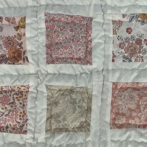 May include: Close-up of a handmade quilt with a patchwork design. The quilt features six fabric squares with floral patterns in shades of pink, peach, and yellow, set against a white background. The squares are framed by white fabric strips.