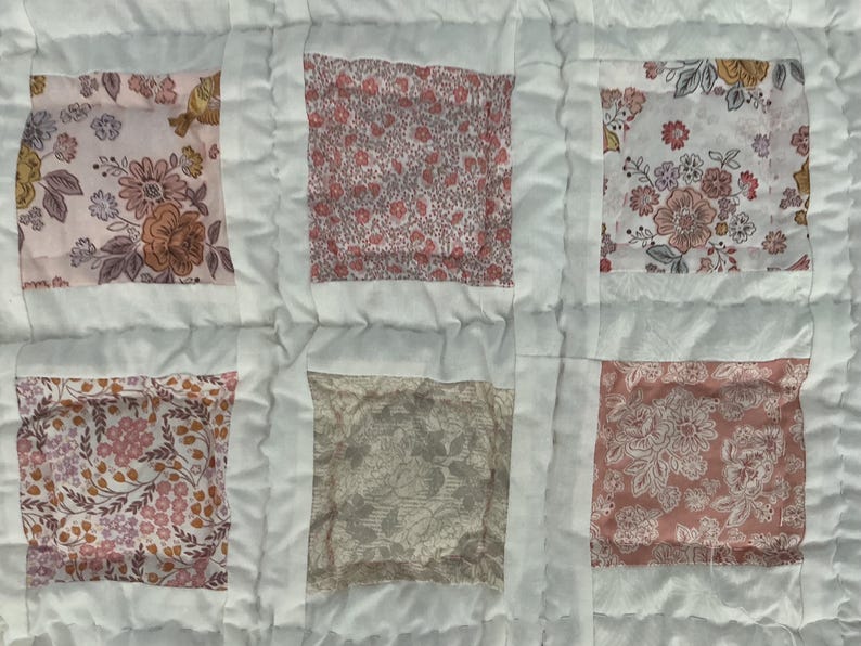 May include: Close-up of a handmade quilt with a patchwork design. The quilt features six fabric squares with floral patterns in shades of pink, peach, and yellow, set against a white background. The squares are framed by white fabric strips.