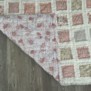 May include: A handmade quilt with a patchwork design. The top features squares in various floral and patterned fabrics in shades of pink, gray, and white. The underside reveals a large floral print in pink and white.