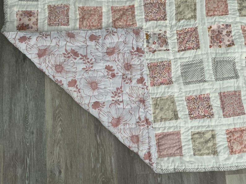 May include: A handmade quilt with a patchwork design. The top features squares in various floral and patterned fabrics in shades of pink, gray, and white. The underside reveals a large floral print in pink and white.