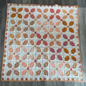 May include: A square patchwork quilt featuring a repeating floral and geometric pattern in shades of peach, coral, rust, and white. The design consists of four-petal flower motifs separated by a white grid, finished with a peach-coloured border.
