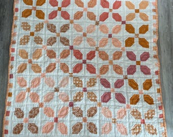 May include: A square patchwork quilt featuring a repeating floral and geometric pattern in shades of peach, coral, rust, and white. The design consists of four-petal flower motifs separated by a white grid, finished with a peach-colored border.