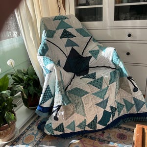 Op de afbeelding: Een doorgestikte deken met een geometrisch patroon van driehoeken in tinten blauwgroen, marineblauw en wit. De stof vertoont diverse texturen en kleine prints, met een centraal donker marineblauw stermotief en een effen marineblauwe rand.