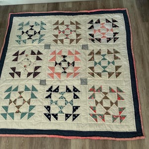 Könnte beinhalten: Eine handgefertigte Steppdecke mit cremefarbenem Hintergrund und neun quadratischen Blöcken. Jeder Block zeigt ein Sternmuster in verschiedenen Farben, darunter Blau, Rosa und Braun. Die Steppdecke ist mit einer marineblauen und rosa Bordüre eingefasst.