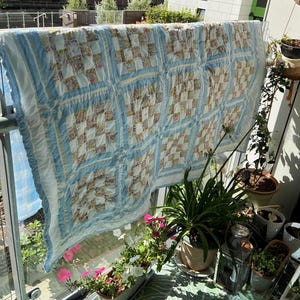 Puede incluir: Un edredón de retazos azul claro y blanco con estampados florales está colgado de una barandilla. El edredón tiene un borde con volantes y está colgado al aire libre en un día soleado. El edredón mide aproximadamente 178 cm de ancho y 203 cm de largo.