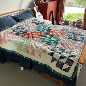 May include: A colourful, handmade quilt with a geometric pattern in white, pink, orange, and teal. The quilt is on a bed with navy blue bedding and a wooden frame. The quilt's design features squares and triangles.
