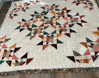 May include: A square quilt featuring a cream-colored background with a repeating star pattern. The stars are constructed from various fabric patches in shades of orange, green, pink, brown, and white. The border has a floral and woodland print.