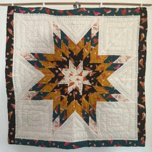 May include: A handmade quilt featuring a star design. The star is crafted from gold, teal, and black patterned fabrics with birds. The quilt has a white background with stitched squares and a dark border with bird motifs.