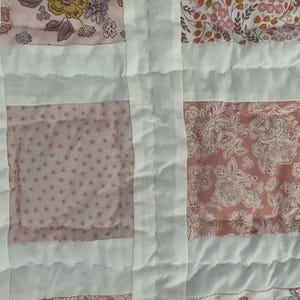 May include: Close-up of a patchwork quilt with a floral pattern. The quilt features squares of pink and white fabric, with a variety of floral designs in shades of pink, peach, and yellow. The squares are separated by white fabric strips.