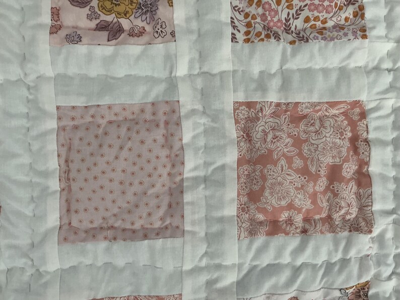 May include: Close-up of a patchwork quilt with a floral pattern. The quilt features squares of pink and white fabric, with a variety of floral designs in shades of pink, peach, and yellow. The squares are separated by white fabric strips.