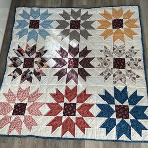 Puede incluir: Una colcha cuadrada con nueve bloques grandes en forma de estrella dispuestos en cuadrícula. Cada estrella tiene un centro granate oscuro y puntas de varios colores, incluyendo azul claro, gris, amarillo, rosa, rojo, azul oscuro y estampados.