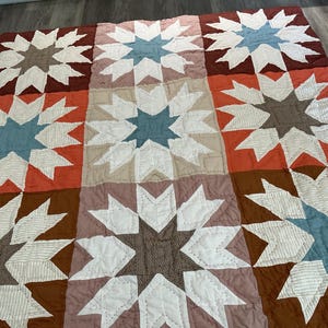 May include: A patchwork quilt featuring a repeating star pattern across nine square blocks. The design incorporates shades of rust, brown, cream, and dusty blue, with visible stitching lines throughout the fabric squares and star points.