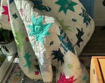 May include: A white quilt featuring a repeating star pattern in various colors including bright pink, teal, lime green, and dark blue with floral and geometric prints. The fabric is stitched with a grid pattern and has a striped edge binding.