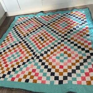 May include: A handmade quilt with a checkerboard pattern in white, teal, pink, navy, and mustard yellow squares. The quilt has a teal border and is lying on a wooden floor.