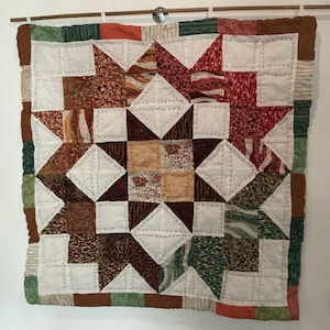 May include: A handmade quilt with a star pattern. The quilt features a central star design with white, brown, red, and green fabric squares. The quilt is hung on a wooden rod with a metal hook.
