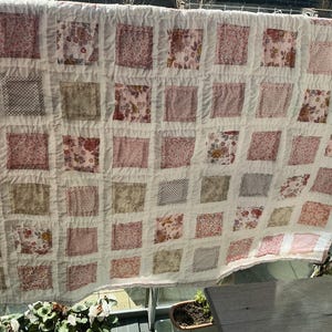 May include: A handmade quilt with a patchwork design. The quilt features squares of various floral and patterned fabrics in shades of pink, red, and beige, set against a white background. The quilt is hanging outdoors.