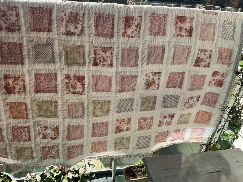 May include: A handmade quilt with a patchwork design. The quilt features squares of various floral and patterned fabrics in shades of pink, red, and beige, set against a white background. The quilt is hanging outdoors.
