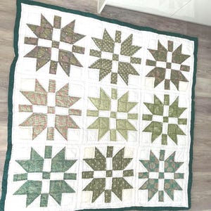 May include: A square quilt featuring a grid of nine star-shaped blocks. Each block is constructed from white fabric and various shades of green patterned fabric. The entire quilt is bordered by a solid dark green fabric binding along the edges.
