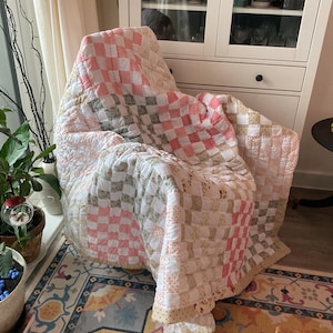 以下が含まれることがあります： ピンク、白、緑のチェック柄のキルトが椅子にかけられています。キルトは花柄で、柔らかくキルティングされた生地で作られています。