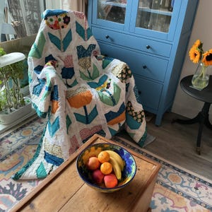 May include: A handmade patchwork quilt featuring a repeating tulip pattern in various colours including orange, blue, green, and floral prints on a white background, draped over a chair. A ceramic bowl filled with oranges and bananas sits nearby.