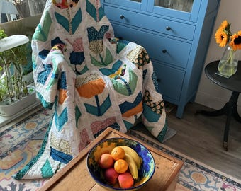May include: A handmade patchwork quilt featuring a repeating tulip pattern in various colors including orange, blue, green, and floral prints on a white background, draped over a chair. A ceramic bowl filled with oranges and bananas sits nearby.