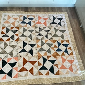 Puede incluir: Un edredón hecho a mano con un diseño geométrico de estrella. El edredón presenta triángulos en tonos blanco, azul marino, melocotón, marrón y gris. El edredón está bordeado con una tela floral color crema y está colocado sobre un suelo de madera.