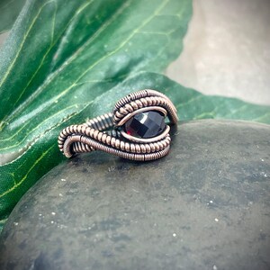May include: A handcrafted ring with a dark red gemstone, intricately wrapped in copper wire. The ring is set on a dark gray stone, with a green leaf in the background. The wirework creates a textured, artistic design.