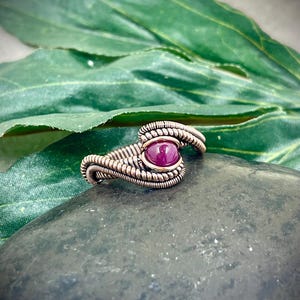May include: A handcrafted wire-wrapped ring featuring a round, deep red gemstone. The ring's intricate design showcases a woven metal band in a copper tone, creating a unique and artistic piece of jewelry. The ring is set on a dark gray stone against a green leaf.
