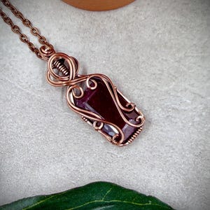 May include: A copper-colored wire-wrapped pendant necklace featuring a rectangular, deep red gemstone. The pendant is suspended from a matching chain. The wirework has intricate swirls and curves, creating an elegant and handcrafted look.