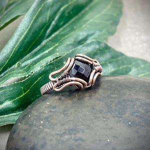 May include: A handcrafted copper wire ring featuring a square, black gemstone. The ring's intricate wirework wraps around the stone, creating a unique and artistic design. The ring is displayed on a dark, textured surface, highlighting its details.