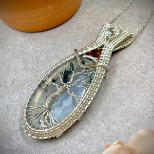 May include: A handcrafted silver pendant necklace featuring a wire-wrapped, oval-shaped stone with a tree of life design. The stone has a blue and green pattern. A small, orange-red stone is set near the top. The pendant hangs from a delicate silver chain.