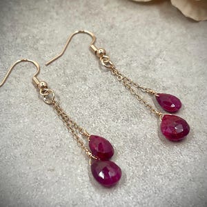 May include: Rose gold earrings featuring two deep red, faceted, teardrop-shaped gemstones suspended on delicate chains. The earrings are displayed on a grey surface. Each gemstone is approximately 1 cm in length.