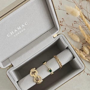 May include: A gray jewelry box with a zipper closure and the text "CHAMAC LONDON" on the lid. The box is open and contains a gold chain bracelet, a gold ring with green stones, and a gold ring with a green stone and clear stones.