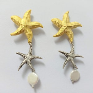 May include: A pair of drop earrings featuring a gold-toned starfish at the top, a silver-toned starfish suspended below, and a small, round, white pearl-like bead dangling at the very bottom of each earring, showcasing a textured surface.