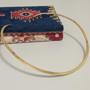 May include: A gold-colored, minimalist collar necklace with a curved design. The necklace rests in front of a stack of two decorative fabric items with a blue and red pattern.