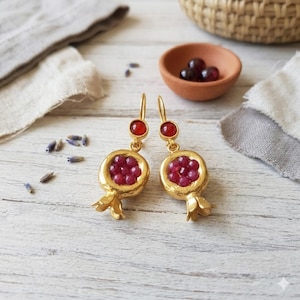 Può includere: Orecchini color oro con design a forma di melograno. Ogni orecchino presenta una gemma rossa rotonda nella parte superiore e un ciondolo a forma di melograno pieno di piccole gemme rosse. Gli orecchini sono esposti su una superficie di legno bianco.
